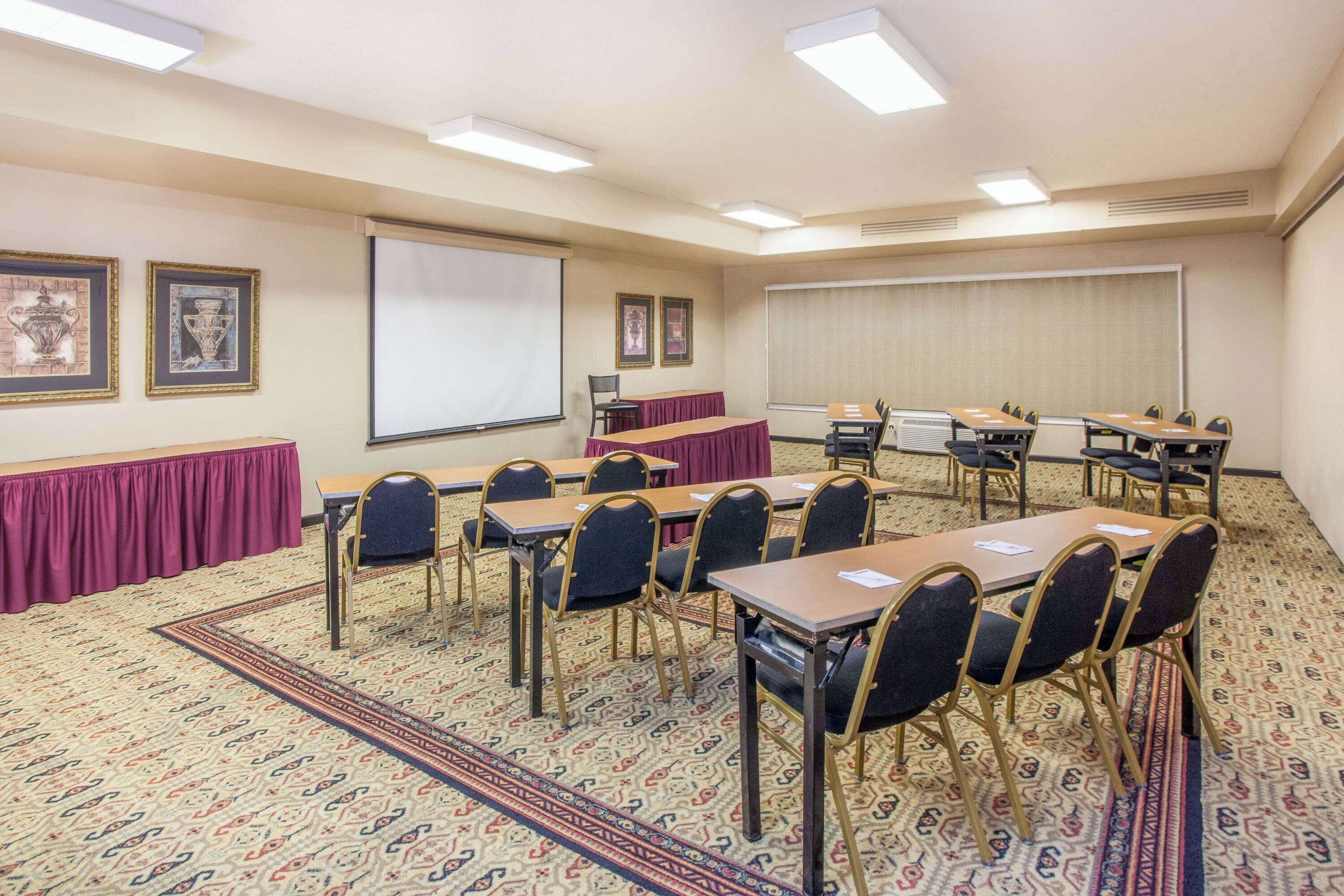 A conference room with a carpeted floor, tables, chairs, a projector screen, and framed pictures on the walls.
