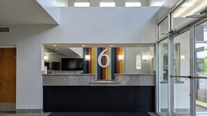 A hotel reception area with a large '6' sign, a reception desk, a flat-screen TV, and a colorful striped carpet. - Studio 6 Pensacola, FL - West I-10 (Pensacola)
