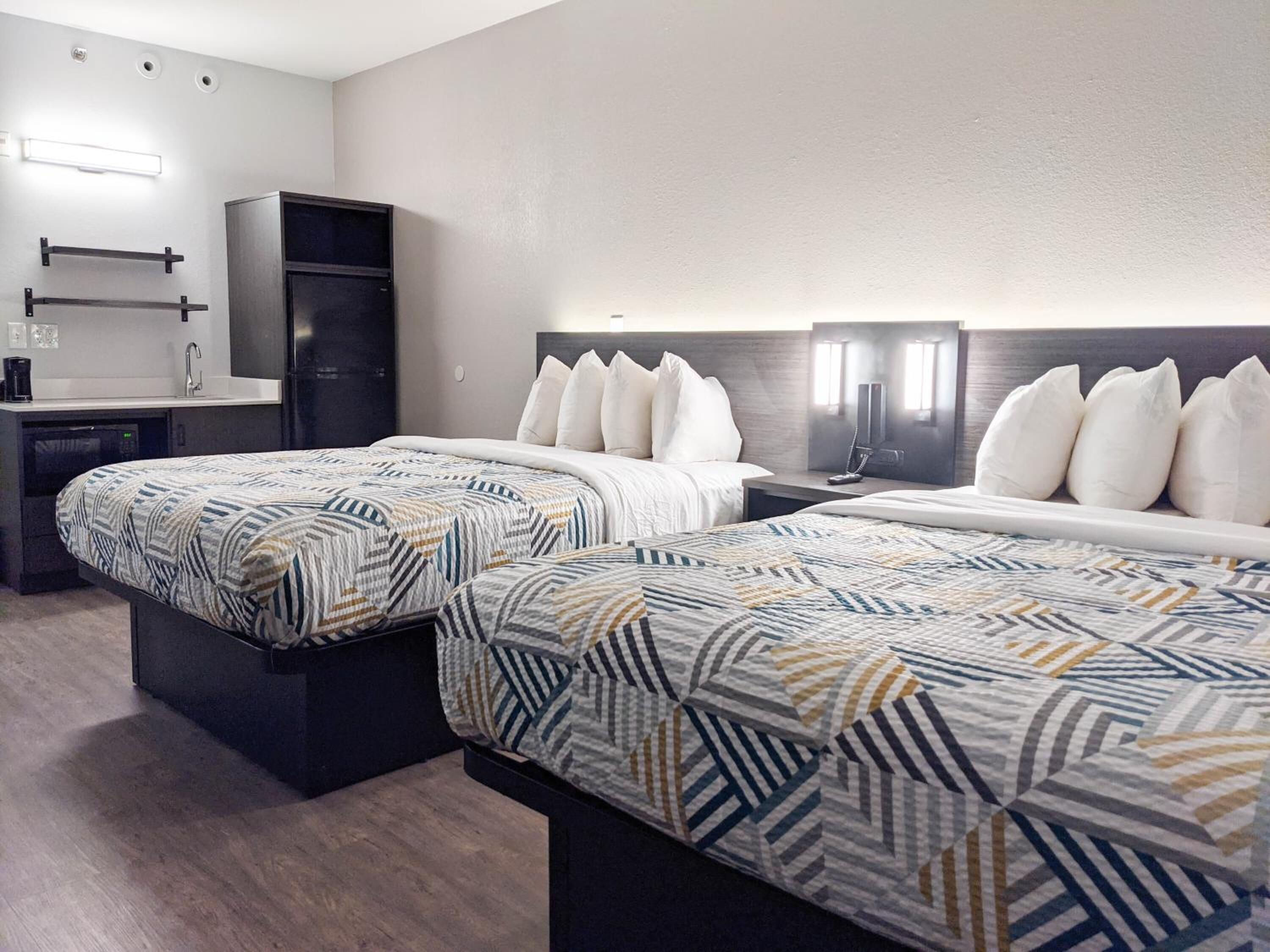 A hotel room with two beds, a small kitchenette, a built-in TV, and a wall-mounted light fixture.