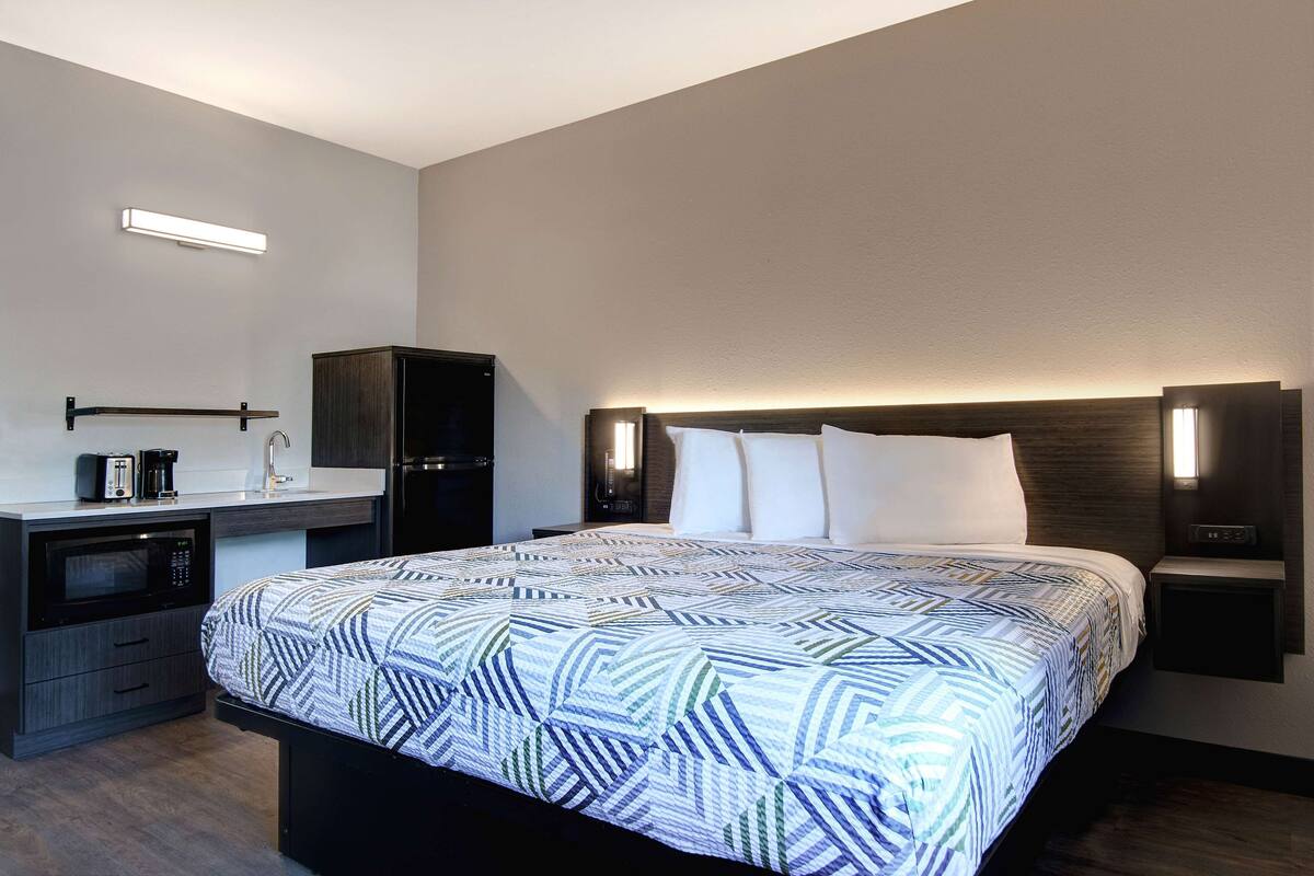A modern hotel room with a large bed, a bedside table with a lamp, a built-in microwave, and a small desk with a coffee maker.
