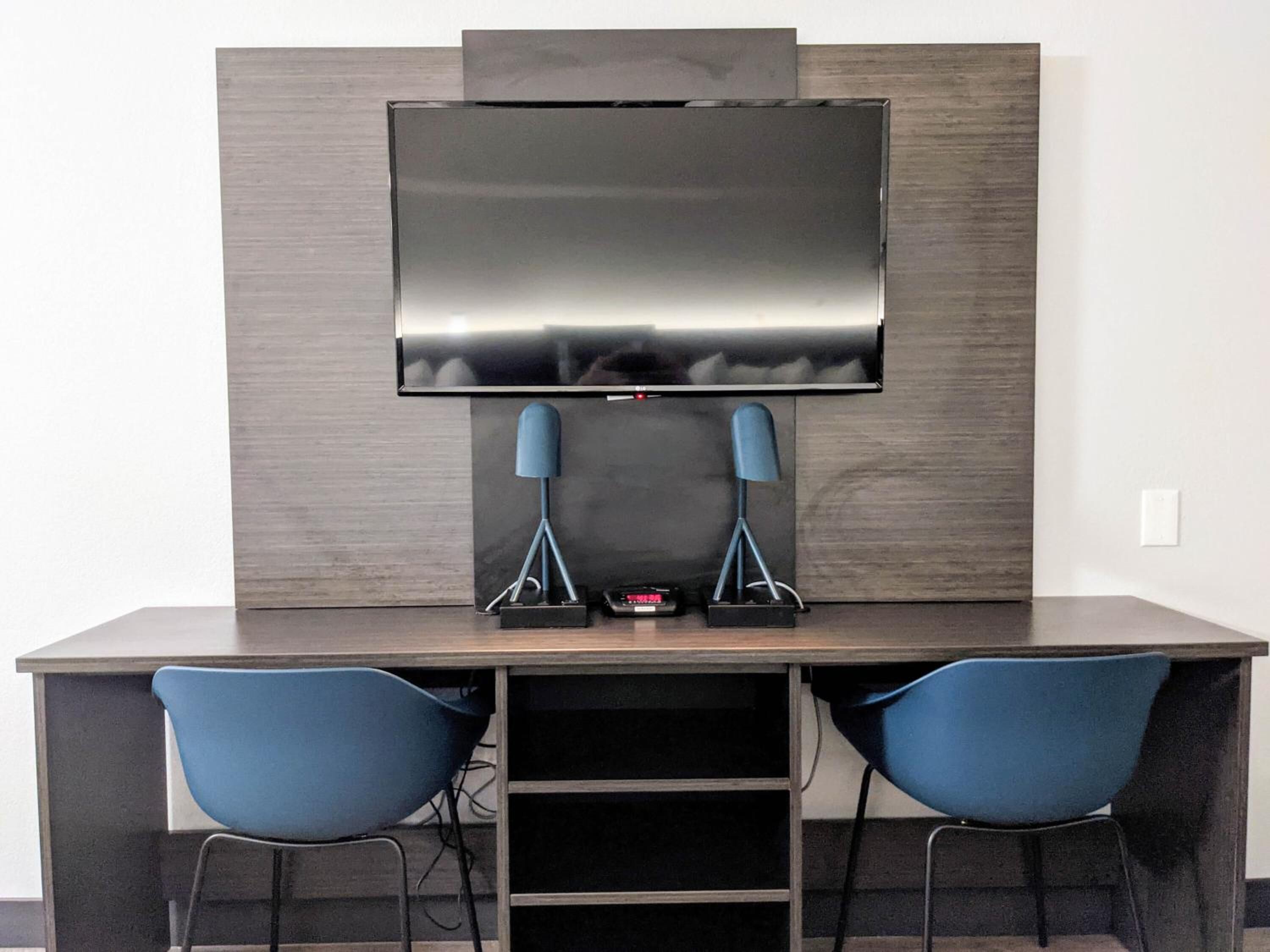 A modern room with a dark wood desk, two blue chairs, a flat-screen TV mounted on the wall, and a small table with a remote control.