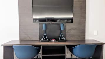 A modern room with a dark wood desk, two blue chairs, a flat-screen TV mounted on the wall, and a small table with a remote control.