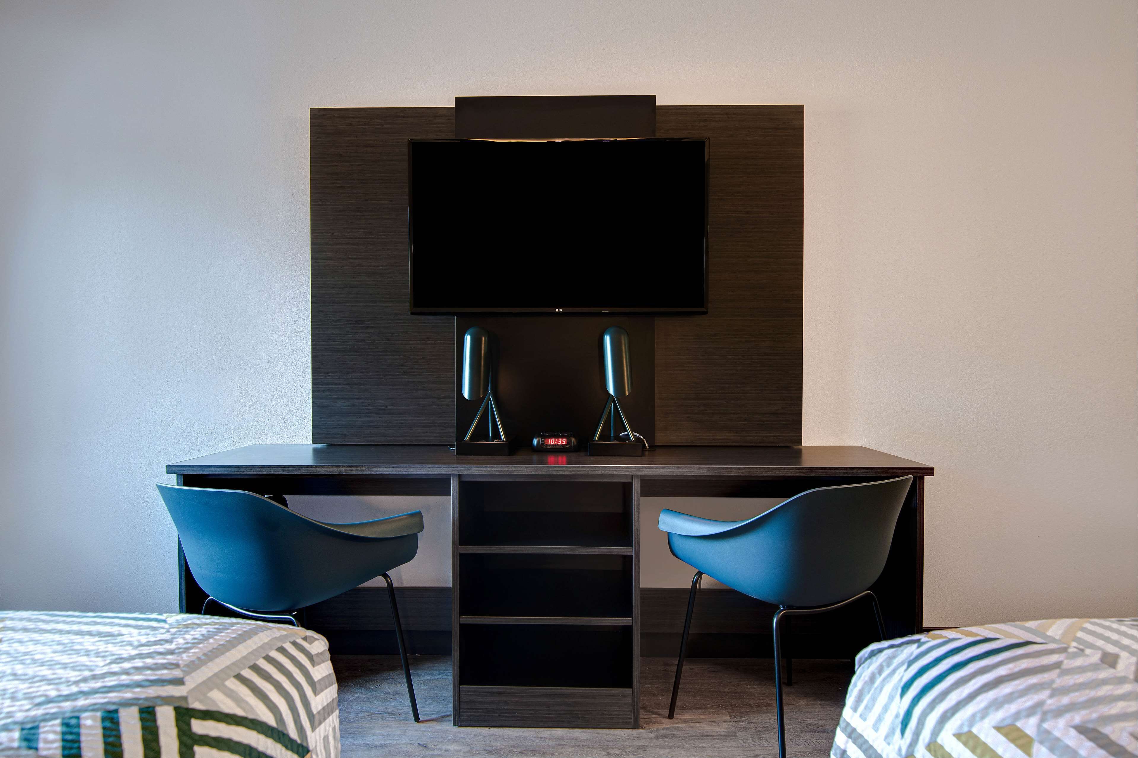 A hotel room with a dark wood desk, two blue chairs, a flat-screen TV mounted on the wall, and a small shelf below the TV.