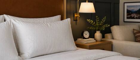 A neatly made bed with white linens, a wooden headboard, a bedside table with a lamp, a clock, and a small plant.