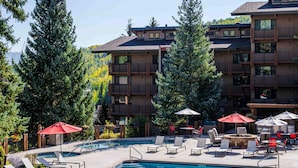 A pool area with lounge chairs and umbrellas, surrounded by tall trees and a multi-story building with balconies. - Stonebridge Inn (Snowmass Village)