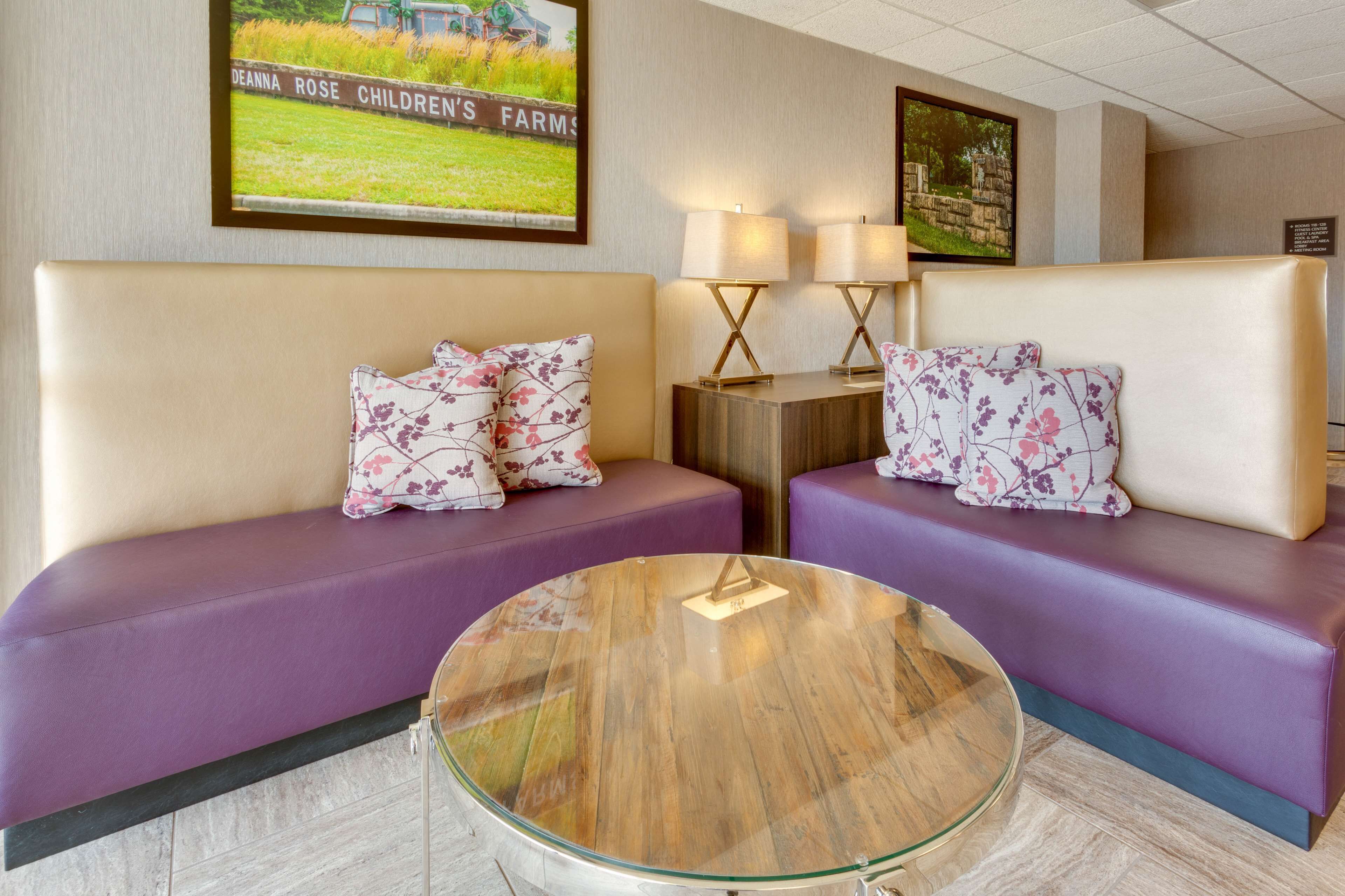 Habitación de hotel con dos sofás morados, una mesa de centro de cristal y una obra de arte en la pared que muestra una escena campestre.