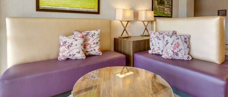 A hotel room with two purple sofas, a glass coffee table, and wall art depicting a farm scene.
