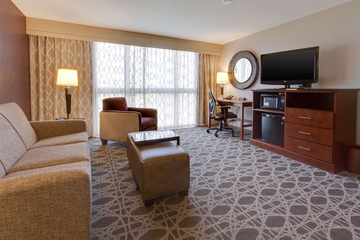A hotel room with a sofa, armchair, ottoman, desk, chair, television, and a microwave.