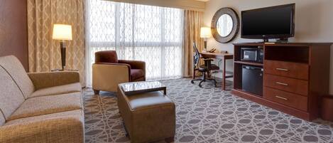 A hotel room with a sofa, armchair, ottoman, desk, chair, television, and a microwave.