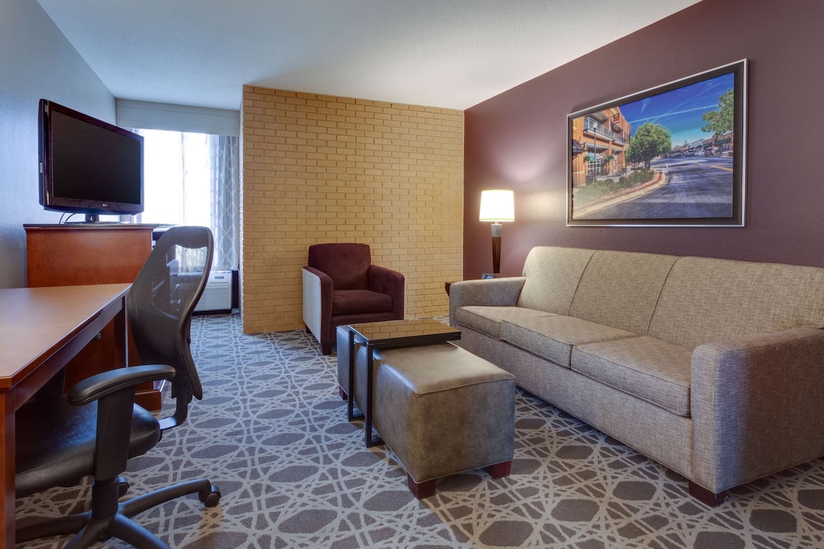A hotel room with a desk, office chair, sofa, ottoman, TV, and a painting on the wall.