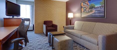A hotel room with a desk, office chair, sofa, ottoman, TV, and a painting on the wall.