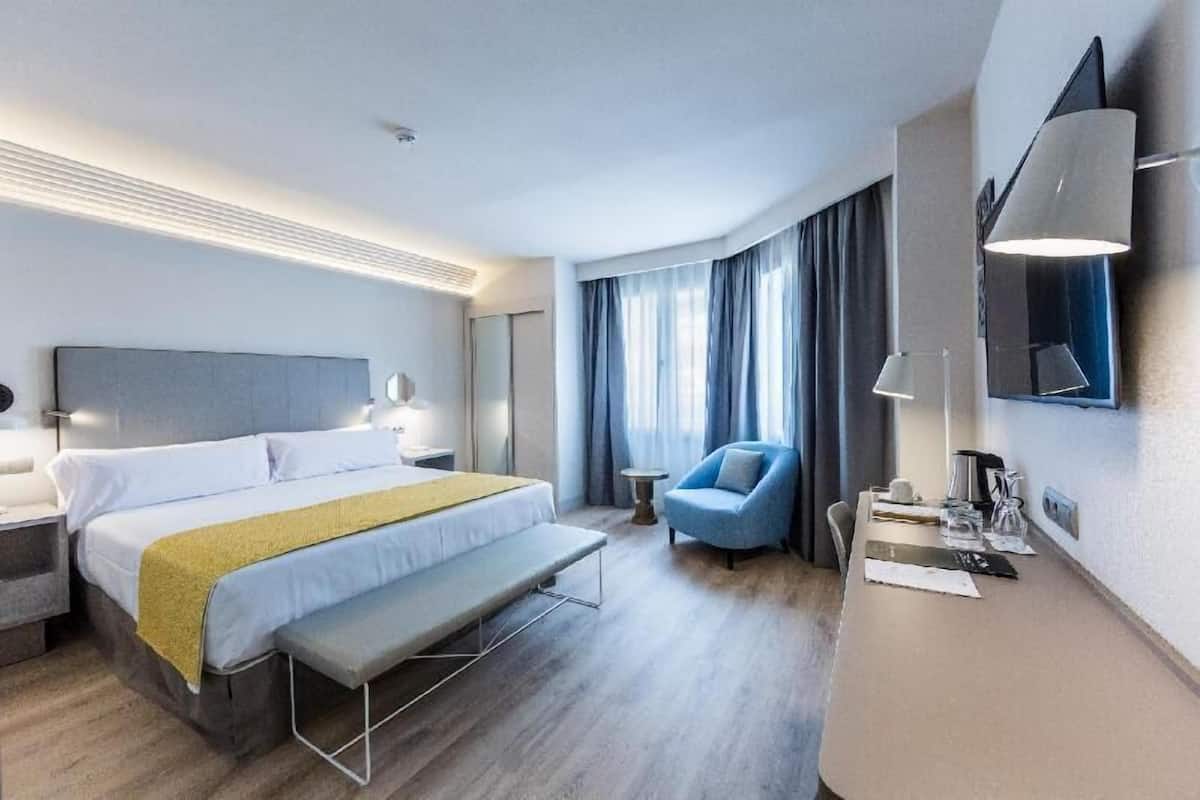 Quarto de hotel moderno com cama, mesa de cabeceira, abajur, cadeira e escrivaninha com luminária.