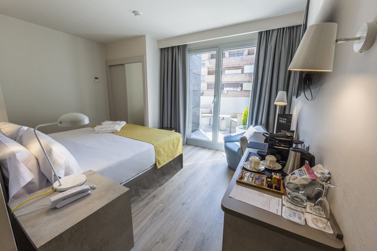 Quarto de hotel com cama, mesa de cabeceira, escrivaninha, cadeira e varanda com vista para edifícios.