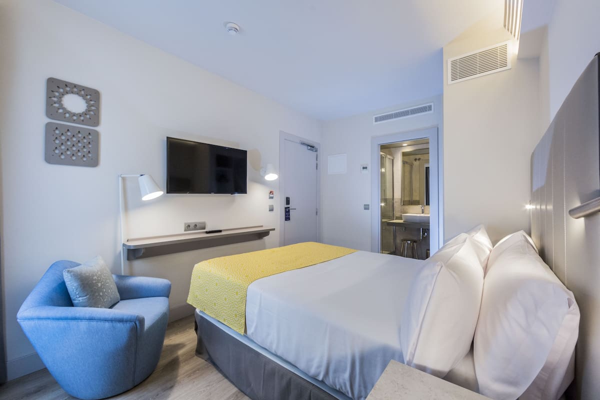 Quarto de hotel moderno com cama, poltrona, televisão e banheiro visível por uma porta aberta.