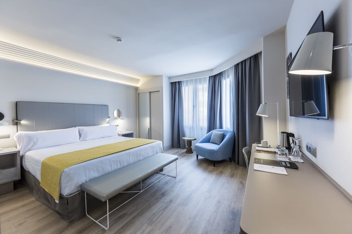 Quarto de hotel moderno com uma cama grande, uma mesinha de cabeceira com abajur, uma pequena mesa redonda, uma poltrona azul e uma escrivaninha com abajur e cafeteira.