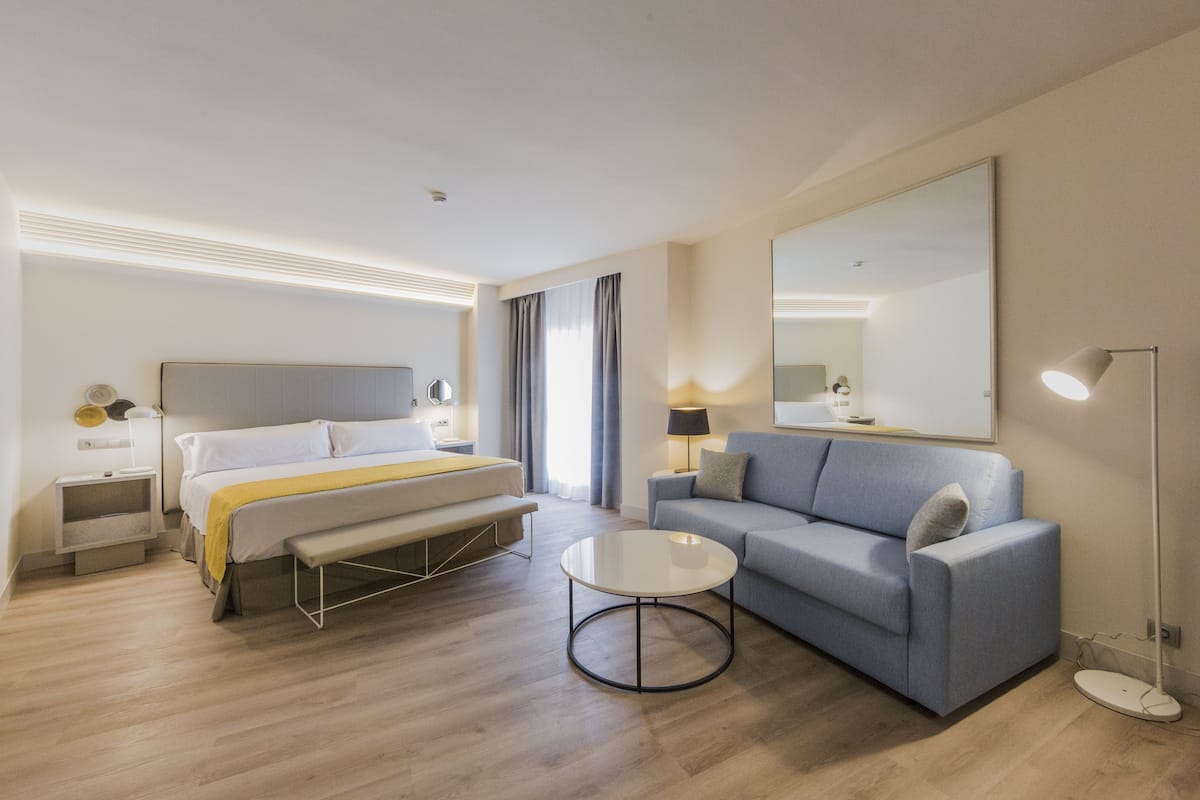 Quarto de hotel moderno com cama, sofá, mesa de cabeceira, luminária e espelho.