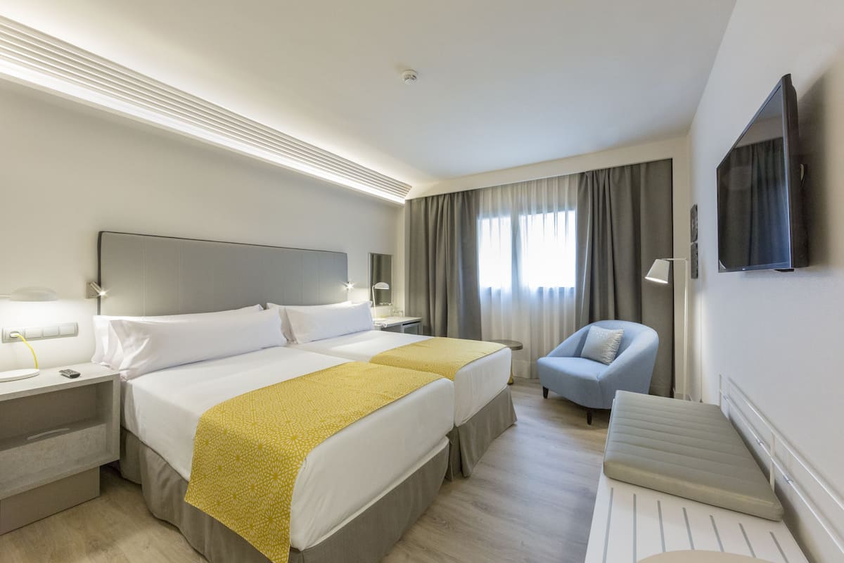 Quarto de hotel com duas camas, uma escrivaninha, uma cadeira, televisão e janela com cortinas.