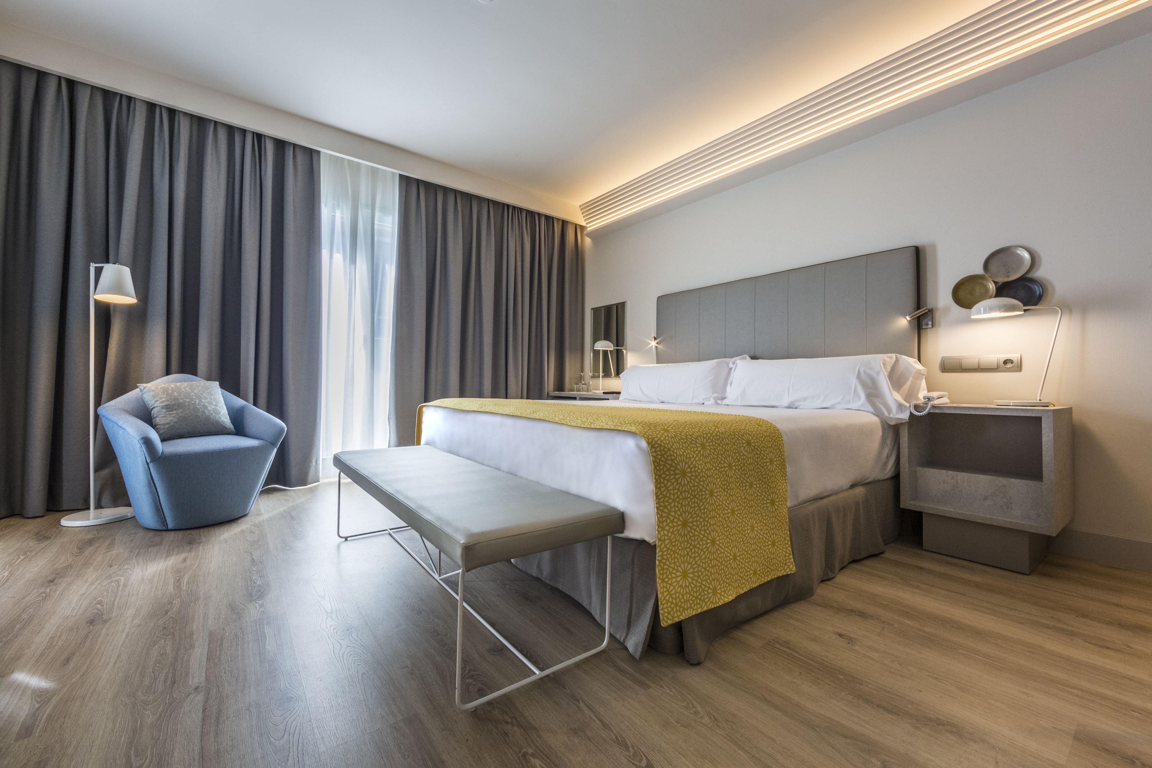 A modern hotel room with a large bed, a bench, a chair, a bedside table, and a nightstand.