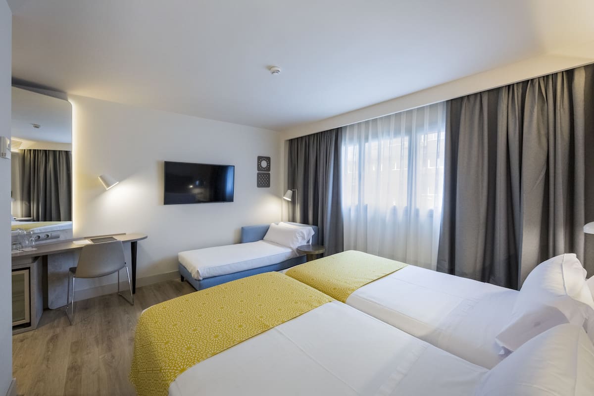 Quarto de hotel com duas camas, uma escrivaninha, uma cadeira, uma televisão e uma janela com cortinas.