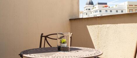 Una mesa metálica redonda con superficie de vidrio, una silla y una pequeña planta en maceta en un balcón con una pared color crema y vista a un edificio con cúpula y estatua.