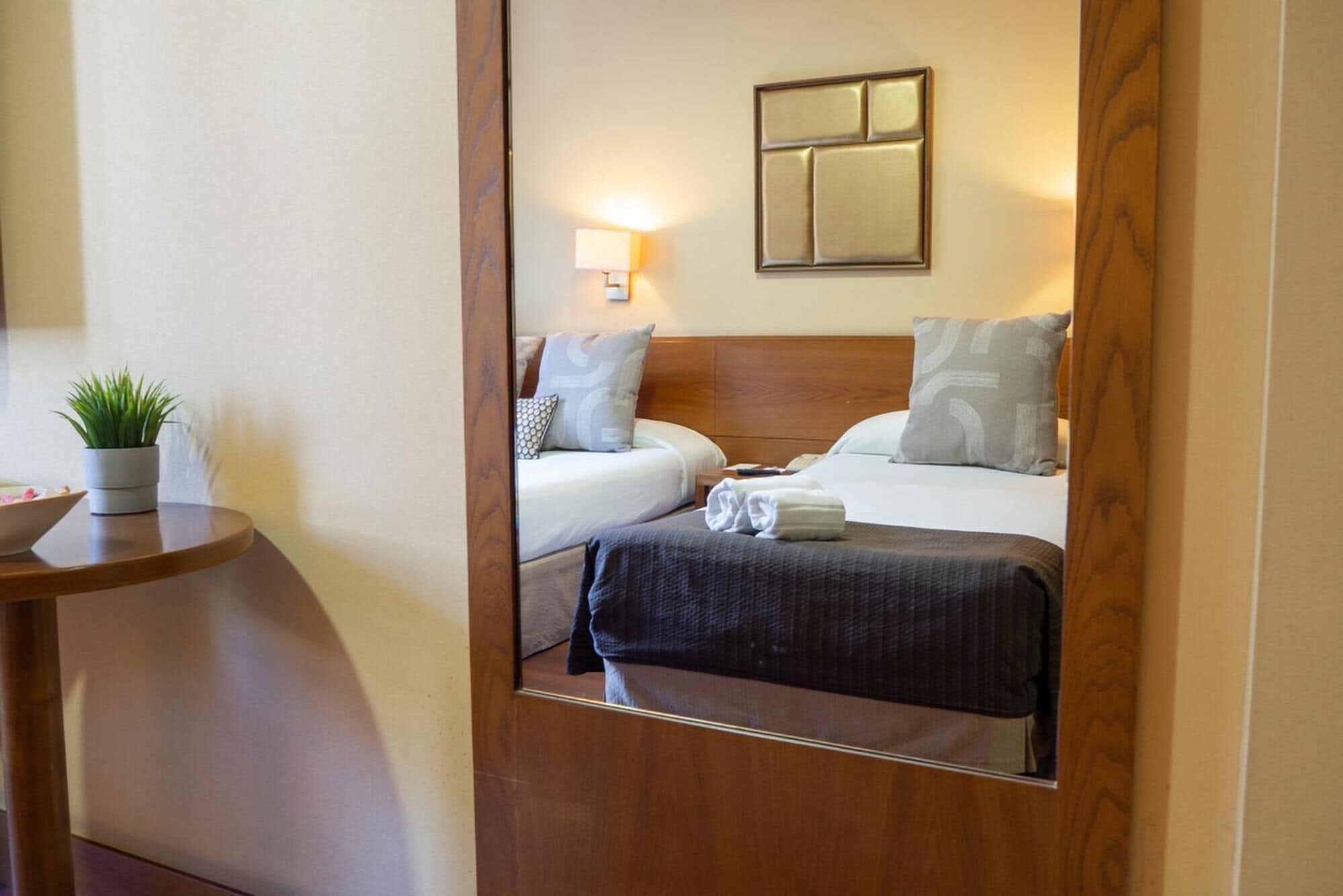 Ein Hotelzimmer mit Bett, Nachttisch, Lampe und einem Spiegel, der den Raum reflektiert.