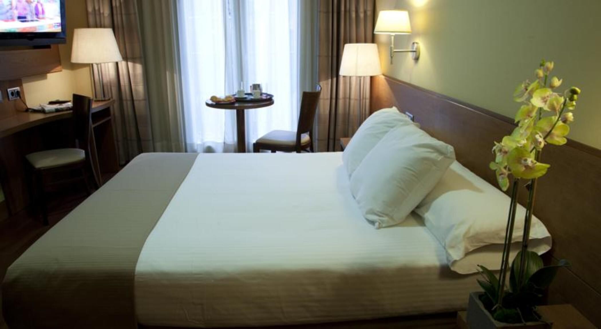 Et hotelværelse med en stor seng, to sengeborde med lamper, et lille bord med stol og et fjernsyn på et skrivebord.