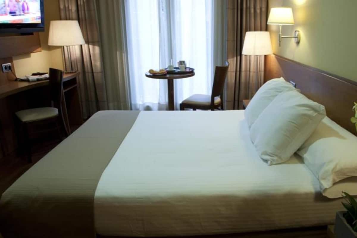 Una habitación de hotel con una cama grande, dos lámparas de noche, una mesita con una silla y un televisor sobre un escritorio.