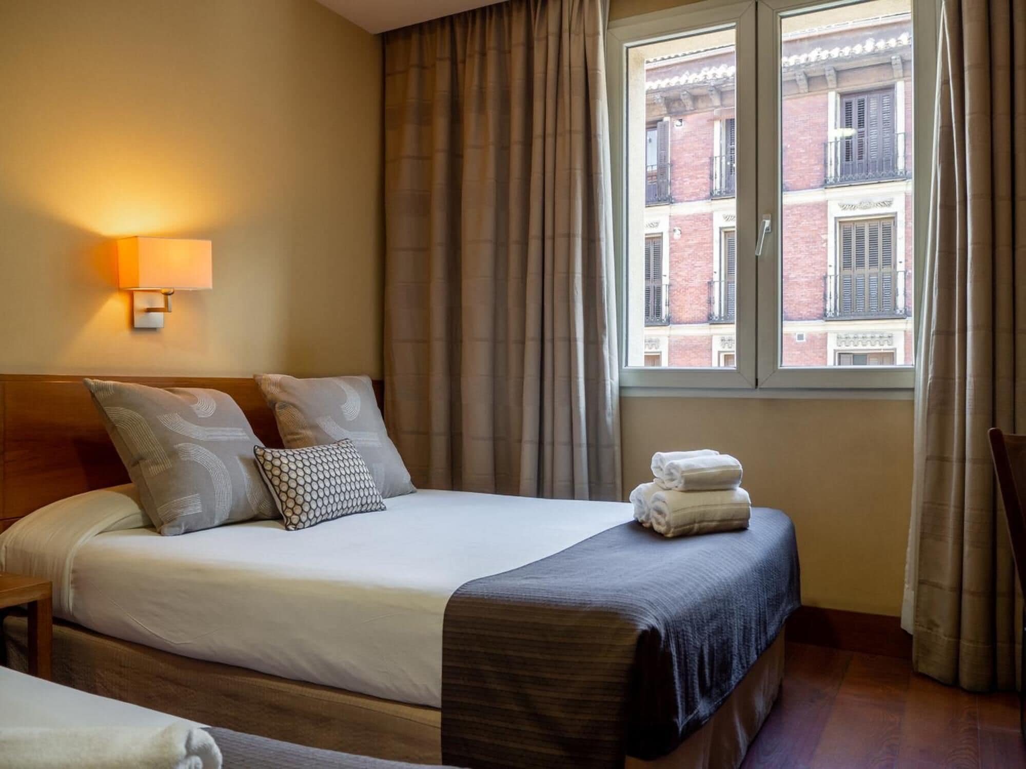 A hotel room with a bed, a bedside lamp, a window with curtains, and a view of buildings outside.