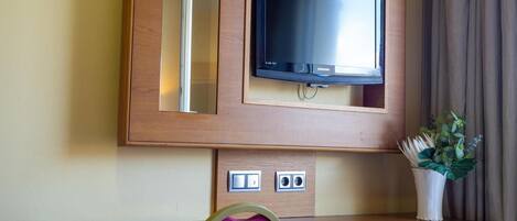 Quarto de hotel com televisão montada, uma mesa com cadeira e uma planta ao lado.