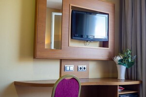 Una camera d'albergo con televisione a parete, una scrivania con una sedia e una pianta.