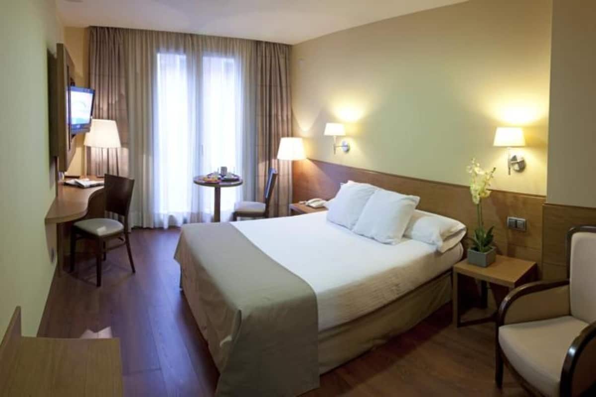 Habitación de hotel con cama, escritorio, silla, una mesita y dos lámparas.