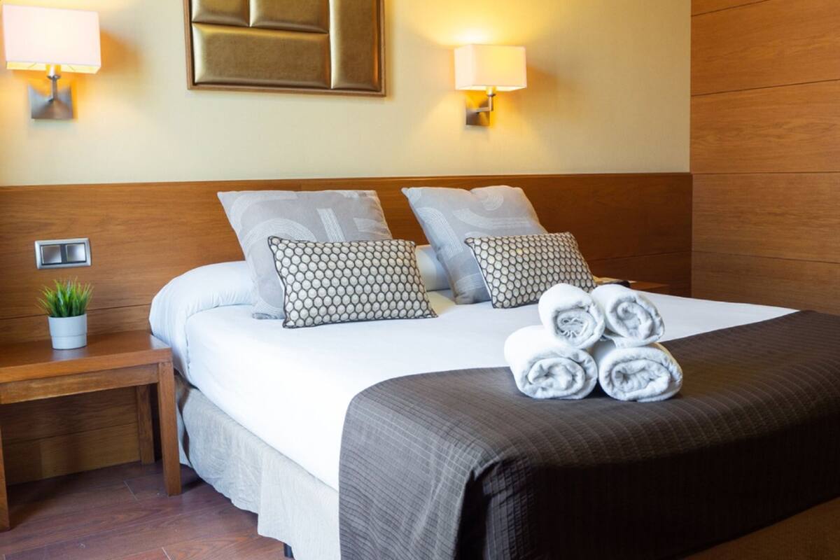 Una cama bien tendida con una manta gris y dos almohadas estampadas, una planta en la mesita de noche y una lámpara de pared.