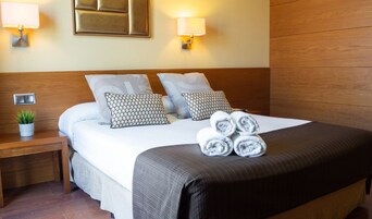 A neatly made bed with a grey blanket and two patterned pillows, a bedside plant, and a wall-mounted light fixture.