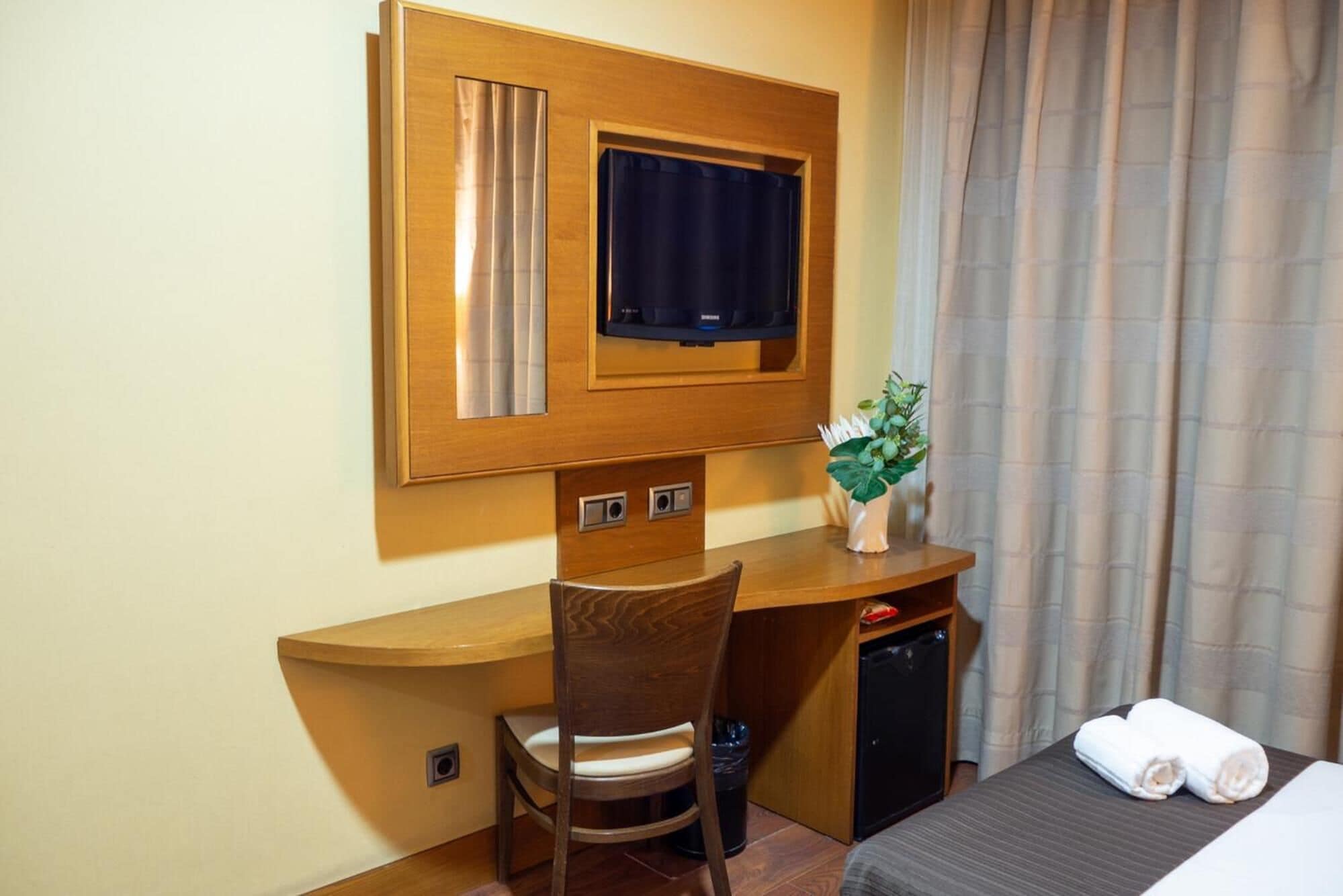 Habitación de hotel con escritorio de madera, una silla, televisor, planta pequeña, cama con una toalla doblada y cortina.
