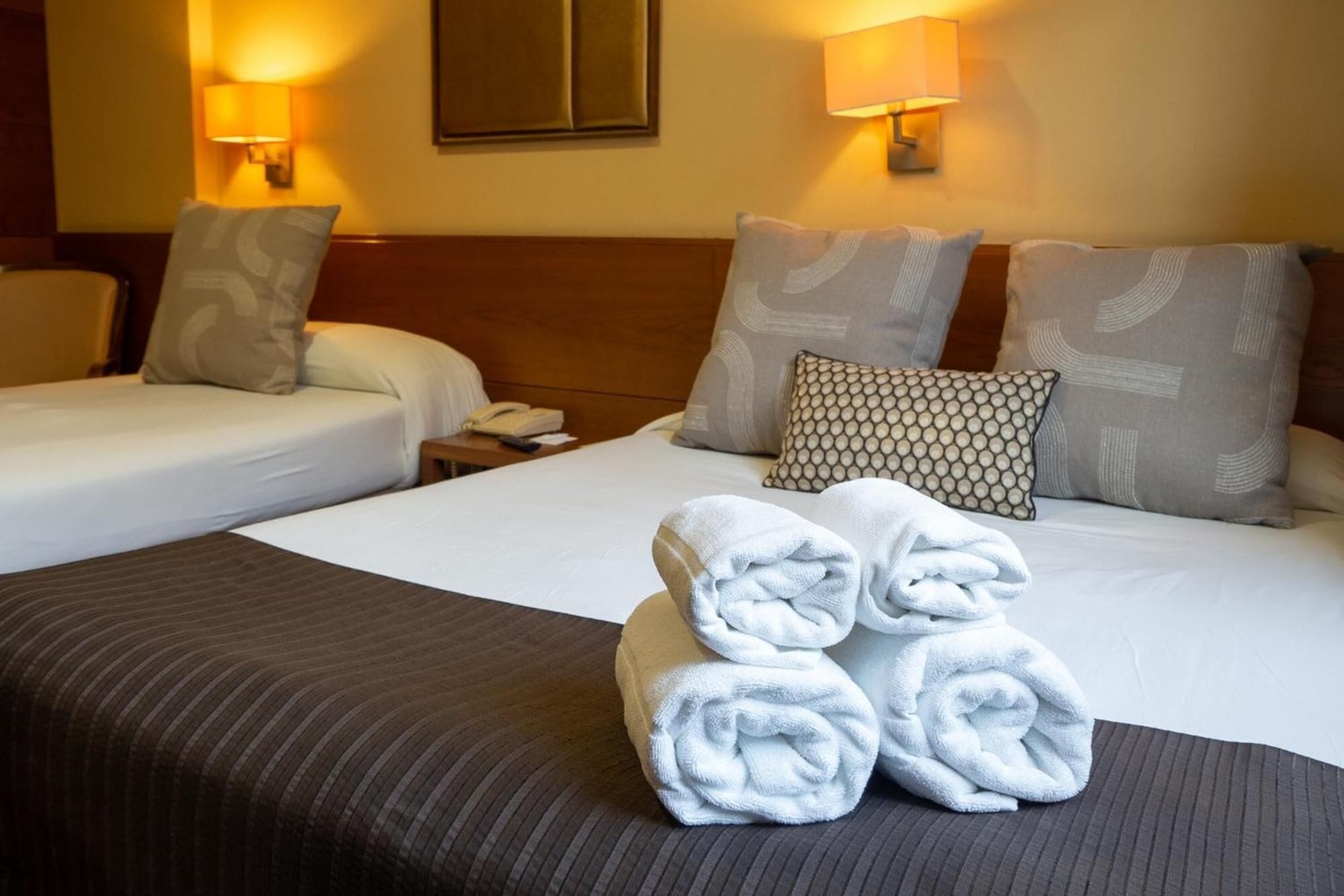 A hotel room with two beds, a bedside table with a phone, and two wall lamps.