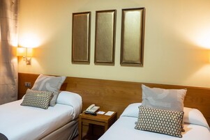 Habitación de hotel con dos camas, cada una con almohada estampada, cabecero de madera, mesita de noche con teléfono y papeles, y tres lámparas fijadas a la pared.
