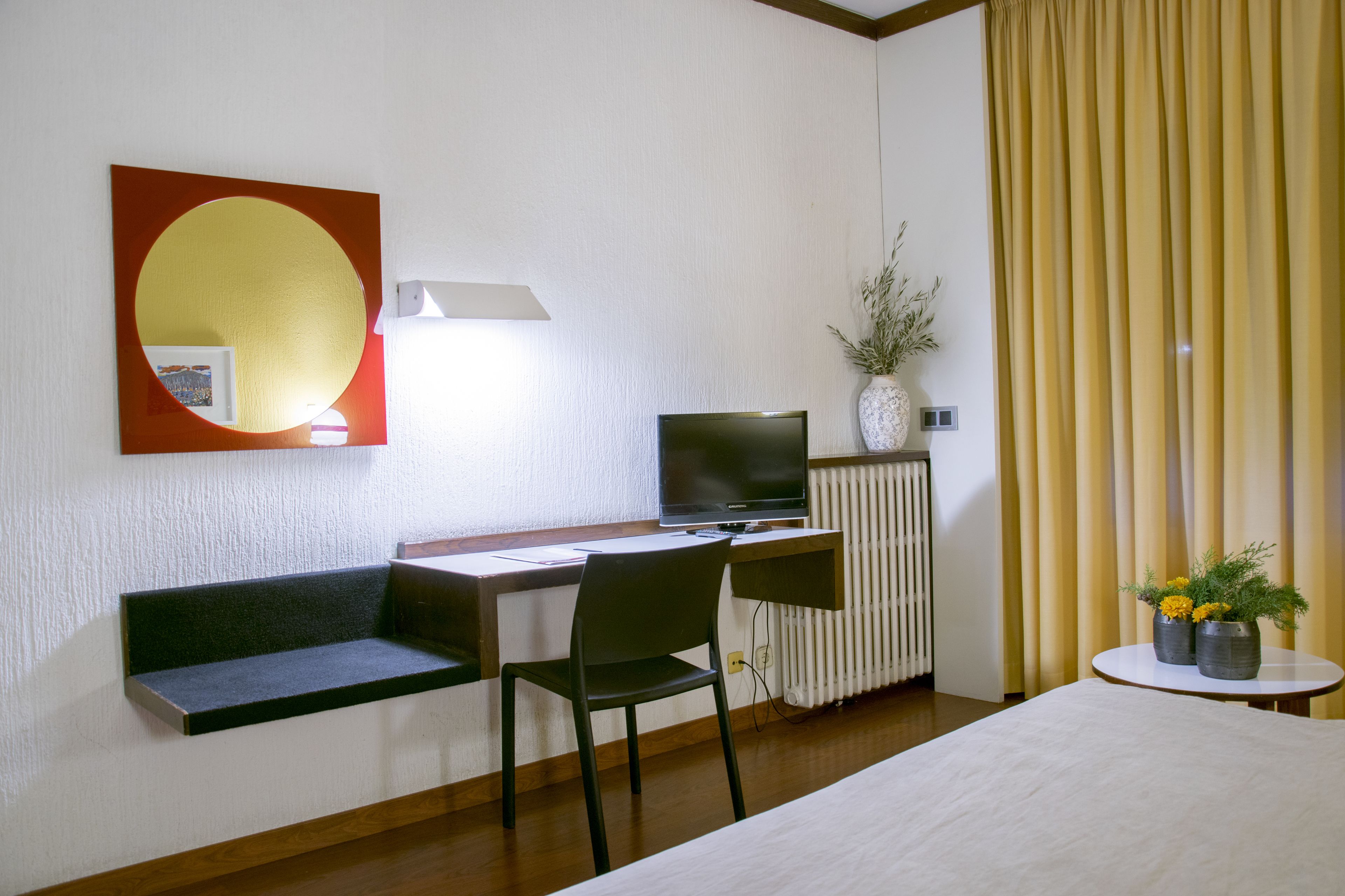 A hotel room with a bed, a desk with a computer, a chair, a small table with a vase, and a window with curtains.