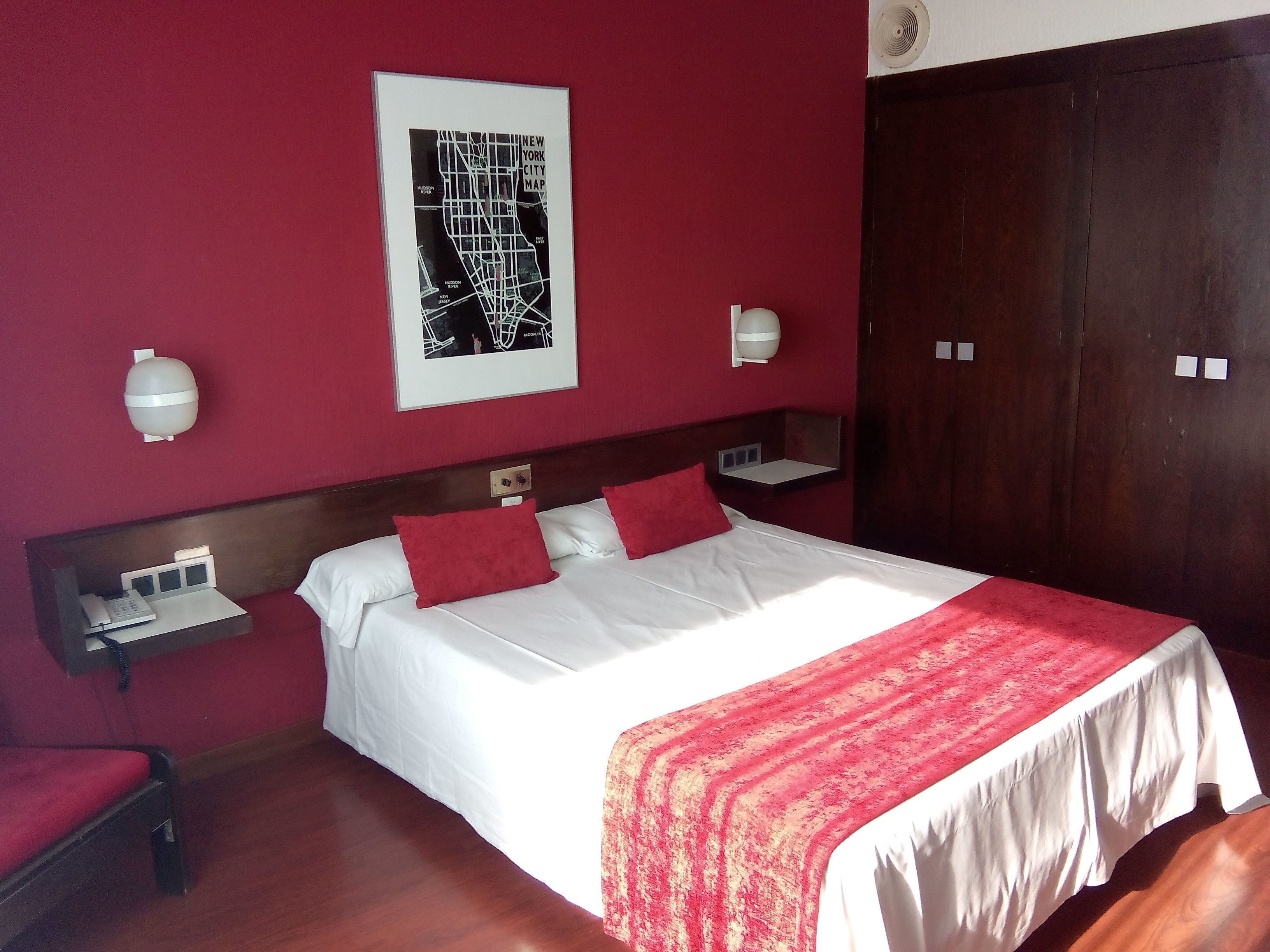 A hotel room with a large bed, red walls, a framed map, bedside lamps, and a wooden wardrobe.