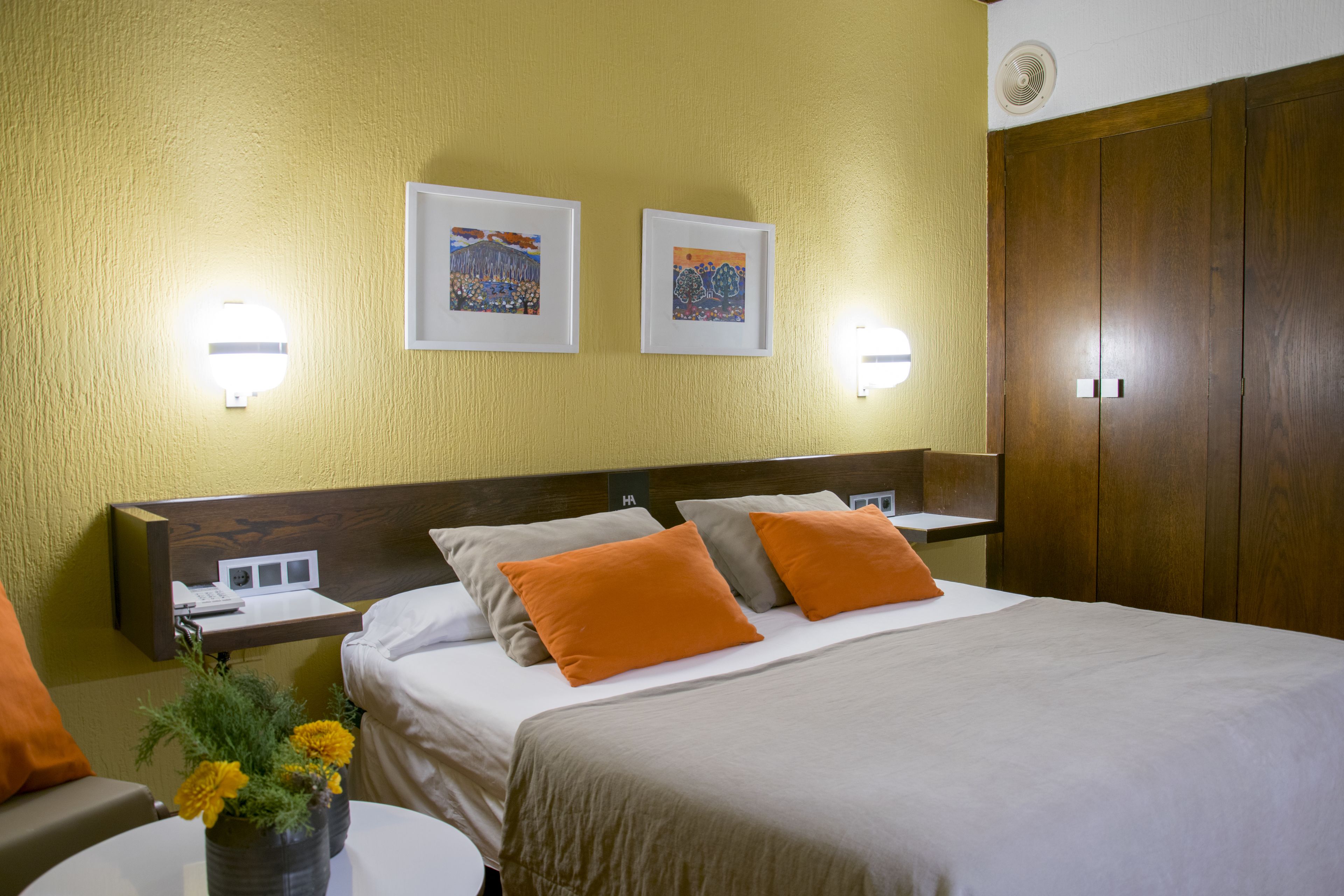 Una habitación de hotel con una cama, dos marcos de cuadros en la pared, una mesita de noche con lámpara y un armario de madera.