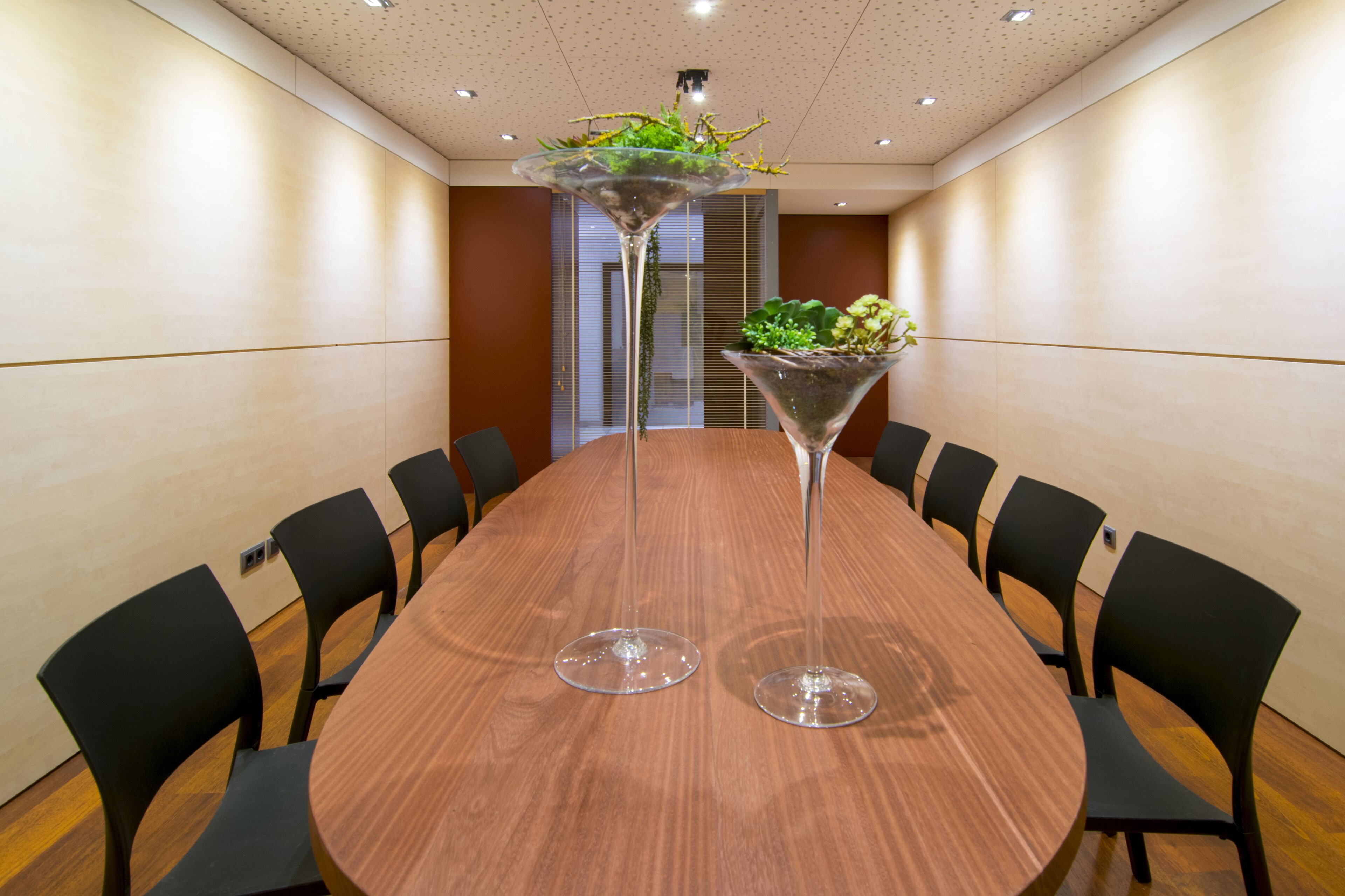 Uma sala de conferência com uma mesa de madeira redonda, cadeiras pretas e dois vasos altos com plantas.