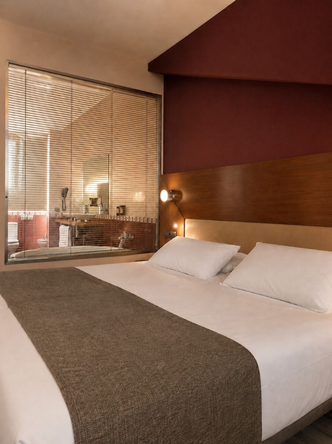 A hotel room with a large bed, a wooden headboard, a large window with blinds, and a view of the bathroom.