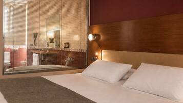 A hotel room with a large bed, a wooden headboard, a large window with blinds, and a view of the bathroom.