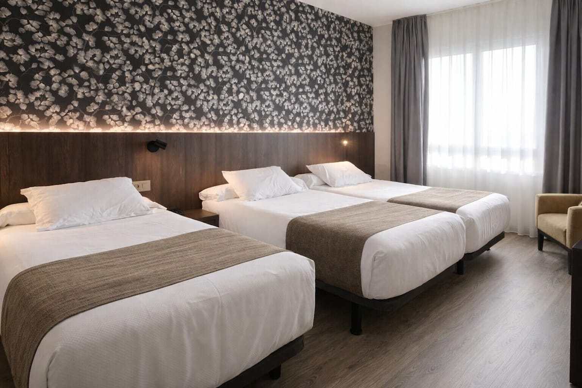 A hotel room with three beds, a wooden headboard, a floral wall design, and a view of the outdoors through a window.