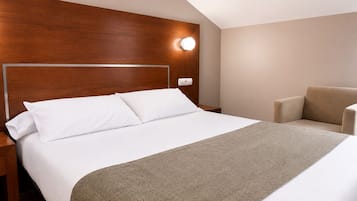 Et hotelværelse med sengegavl i træ, seng med hvide sengetøj, en beige lænestol og vægmonteret lampe.