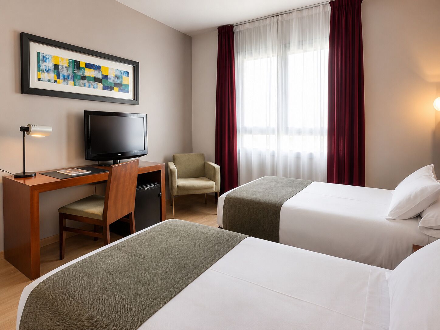 Ein Hotelzimmer mit Schreibtisch, Fernseher, zwei Betten, einem Sessel und einem Fenster mit Vorhängen.