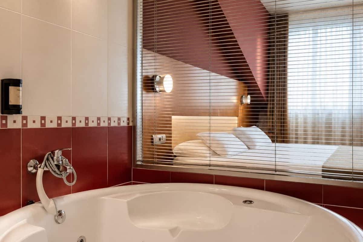 A modern bathroom with a large white bathtub, a shower with a chrome faucet, and a window with blinds reflecting a bedroom interior.