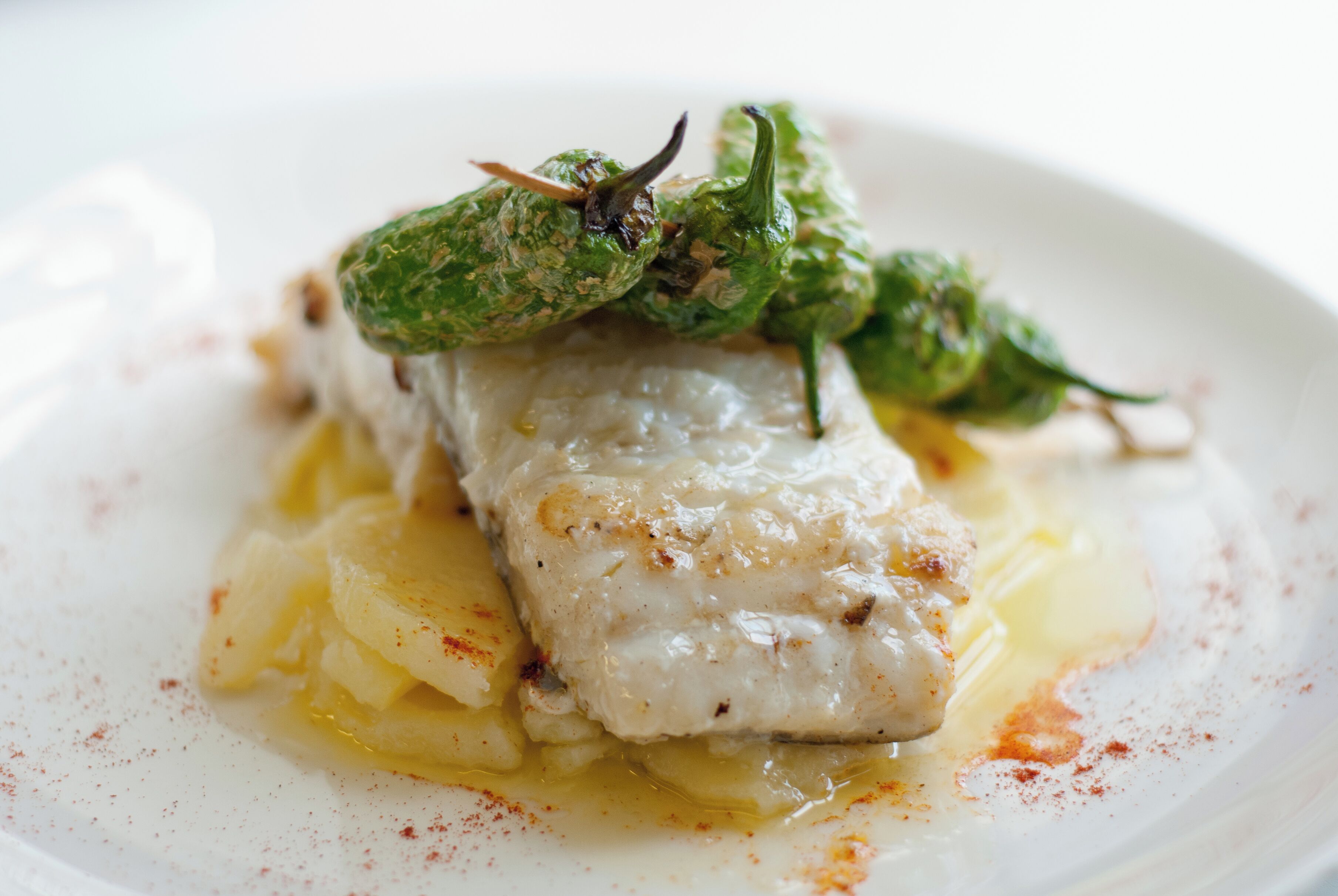 A white plate with a piece of white fish, green chili peppers, and a yellow sauce.