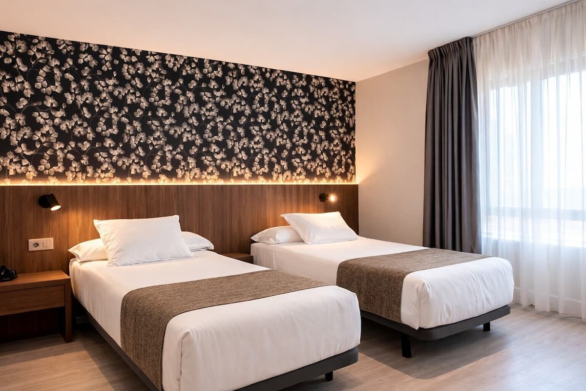 A hotel room with two beds, a wooden headboard, a floral wall design, and a large window with curtains.