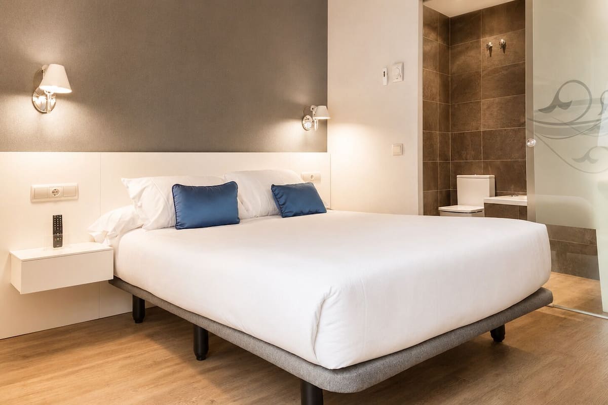 A modern bedroom with a large bed, bedside tables, a nightstand, a wall-mounted lamp, and a bathroom with a shower and toilet.