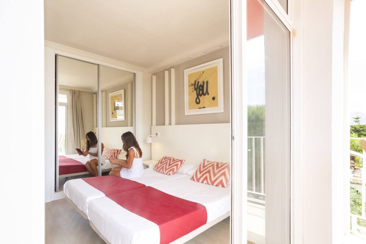 Een slaapkamer met een groot bed, een spiegel, een muur met een ingelijst kunstwerk en een raam met uitzicht op groen.